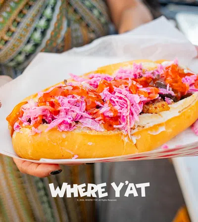 Close-up of a hand holding a hot dog with pink slaw and red sauce in a paper tray, with overlaid text reading WHERE Y'AT.