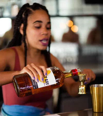 Woman pouring rum from Mount Gay Barbados bottle into a jigger at a bar counter for cocktail preparation