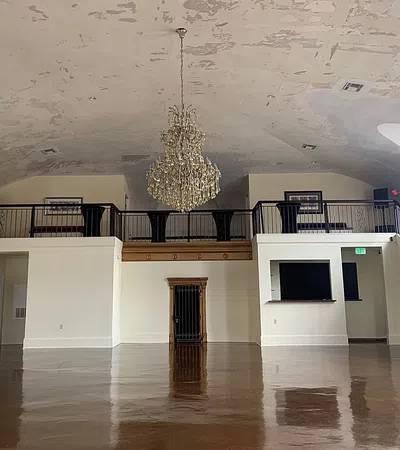 Empty spacious room with polished floor, decorative chandelier, balcony with railings, and peeling ceiling paint.