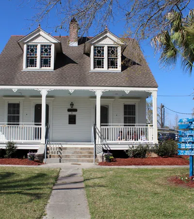 White historic house with a wraparound porch, manicured lawn, and a blue directional sign near a palm tree.
