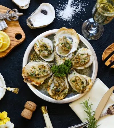 Grilled oysters on plate with lemon, salt, herbs, knife, fork, and glass of white wine on black table.