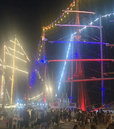 Crowd gathers at night by tall ships decorated with colorful lights and illuminated sails at a festive event.