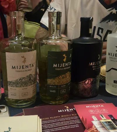 Variety of Mijenta tequila bottles displayed on table with promotional cards and people in the background.