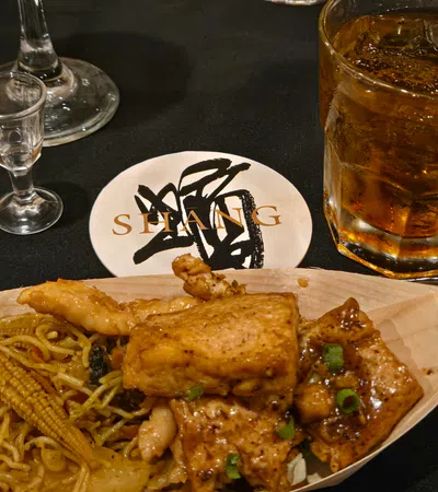 Asian stir-fry with grilled chicken, baby corn, noodles, and iced whiskey on a black table with shot glass.