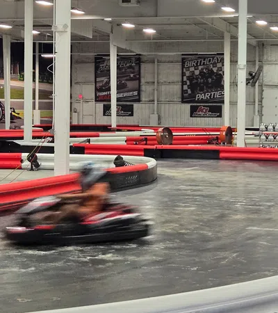 Indoor go-kart racing track with blurred driver speeding around the corner, red and white barriers, and racing posters.