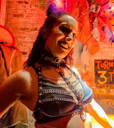 Woman in black leather harness and chains smiles against a brick wall with Halloween decor and overlaid text sign.
