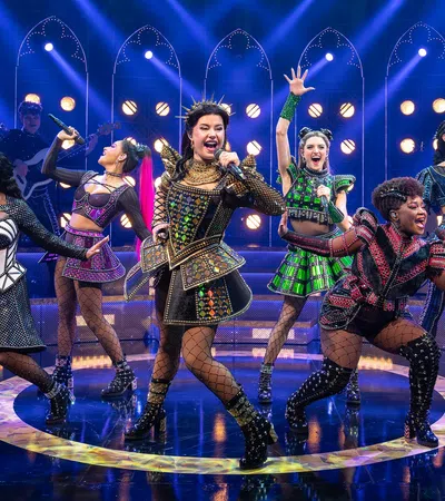 Six female performers sing and dance energetically on a brightly lit stage with colorful costumes and fishnet stockings.