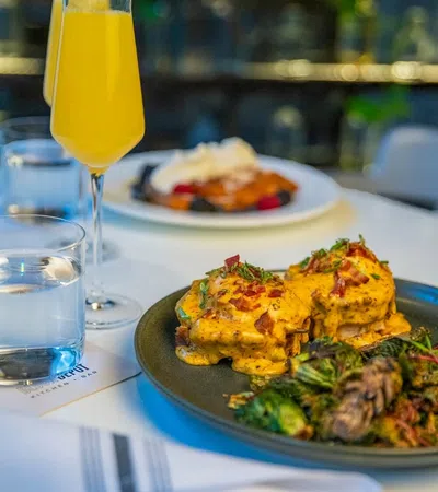 Brunch plate with eggs Benedict topped with hollandaise sauce and crispy bacon, served with sautéed greens and a mimosa.