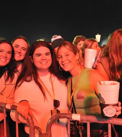 Four smiling young women posing behind a metal barrier at a nighttime outdoor event or concert, holding drinks.