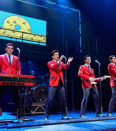 Four male performers in red jackets play instruments and sing on stage with musical notes backdrop in blue lighting.