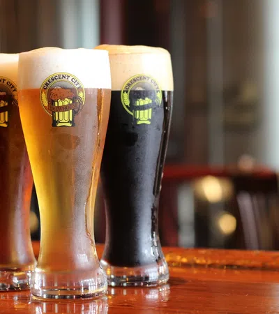 Three tall glasses of Crescent City Brewery beer with varying colors and foamy heads on a wooden bar counter.