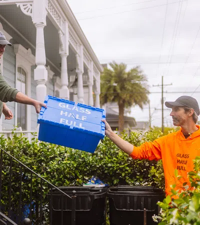 Two men exchanging a blue recycling bin labeled GLASS HALF FULL in a residential outdoor setting with greenery.