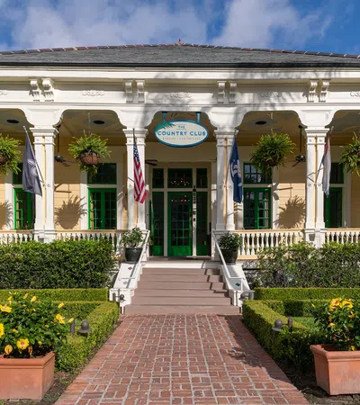Historic country club building with white columns, hanging plants, flags, and well-manicured gardens on a sunny day