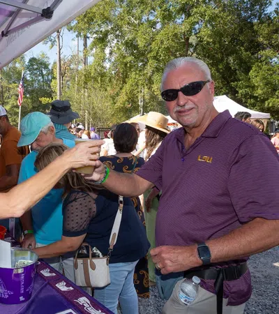 People enjoying a sunny outdoor festival, sharing drinks under a tent with vibrant purple banners.