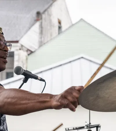Man wearing a beret playing drums energetically at an outdoor performance with houses in the background