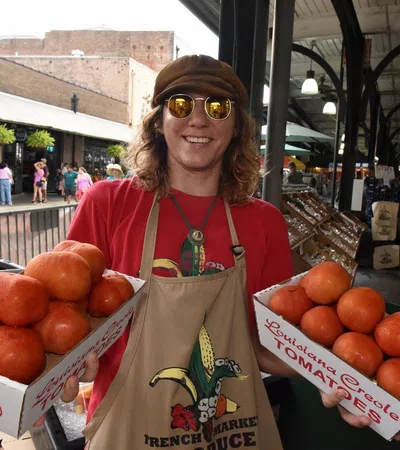 Smiling vendor wearing sunglasses and an apron holds two boxes of fresh Louisiana Creole tomatoes at a busy market.