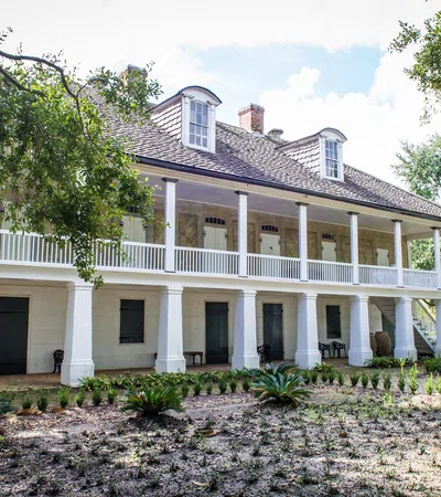 Historic two-story plantation house with white columns, balcony, lush greenery, and a sunny sky background