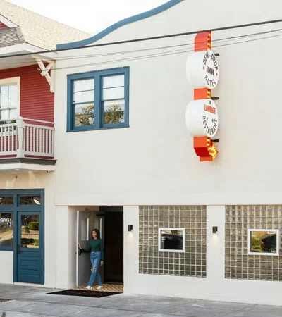 Modern white building with blue accents and a red house with a balcony on the left side in a sunny street.