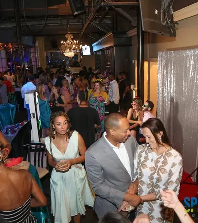 Crowded lively indoor party with guests socializing, dressed in formal and cocktail attire under chandeliers.