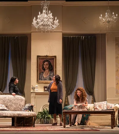 Theatrical stage scene with five women in vintage living room setting under chandeliers and art-adorned walls