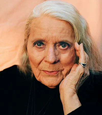 Elderly woman with silver hair, wearing black, resting her face on her hand against a soft peach background.