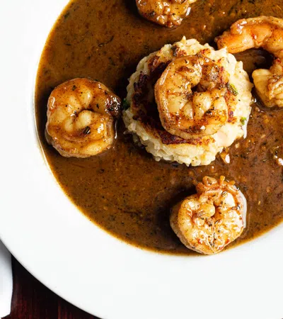 Spicy shrimp served over creamy grits with rich brown sauce on a white plate beside vintage silverware.