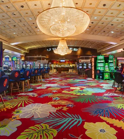 Colorful casino interior with floral carpet, gaming machines, blue chairs, and large chandeliers overhead