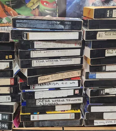 Stack of vintage VHS tapes with handwritten labels on a cluttered desk surrounded by posters and household items.