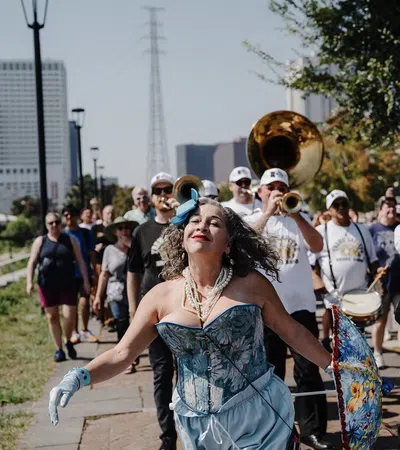 Woman in blue corset and gloves dances joyfully in front of a marching band and parade crowd on sunny day.