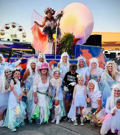 Group of people in white costumes and wings posing outdoors at a festival with a ferris wheel and colorful float.
