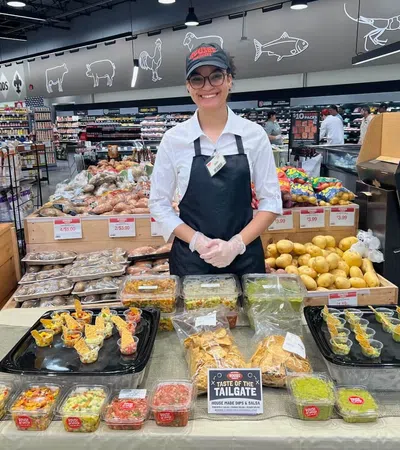 Smiling store employee presenting various homemade dips and salsas at a grocery store sampling table.
