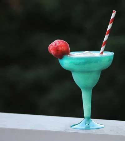 Blue tropical cocktail in a glass with ice, garnished with a red fruit and striped straw, on a white ledge.