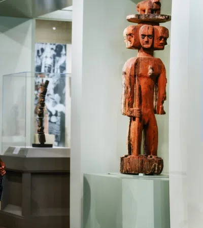 Two museum visitors observe tall wooden tribal sculptures in a brightly lit gallery space with displayed artifacts.