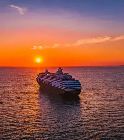 Large cruise ship sailing on calm ocean waters during vibrant orange sunset with clear sky.