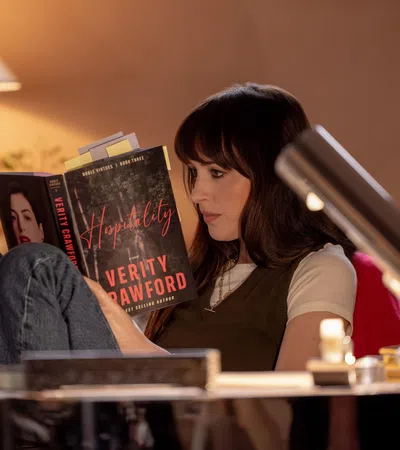 Woman reading the book Hospitality by Verity Crawford in a cozy, warmly lit room with a table lamp.