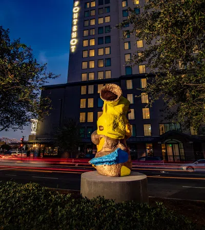 Yellow and blue abstract sculpture on a city street at night with building and light trails in background