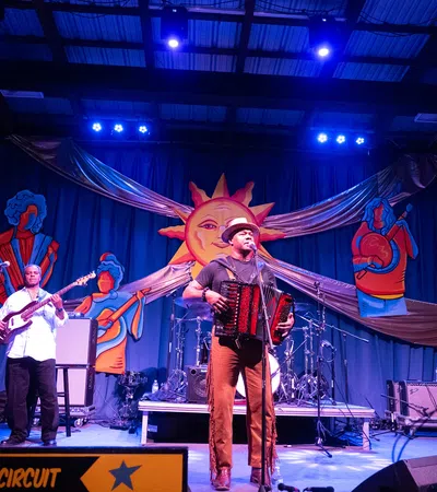 Live band performing on stage with accordion, guitar, and bass under bright lights and colorful sun backdrop