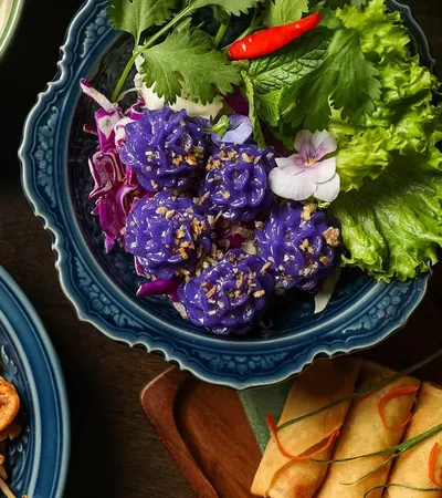 Colorful Thai dishes including purple flower-shaped dumplings, spring rolls, pad thai, and curry on ornate plates.