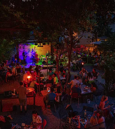 Outdoor evening concert with people seated at tables, colorful stage lights, and festive string lights among trees.