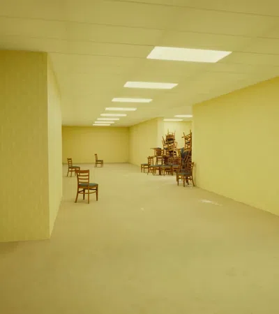 Empty beige room with chairs sparsely placed and a large stack of chairs in the corner under ceiling lights