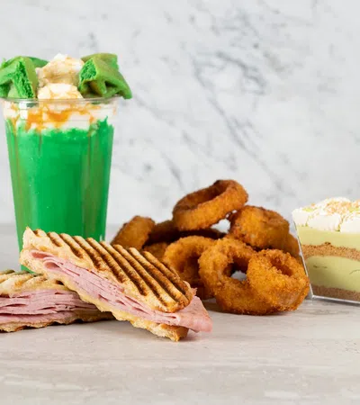 Grilled ham sandwich with fried onion rings, green milkshake topped with whipped cream, and layered dessert on marble table.