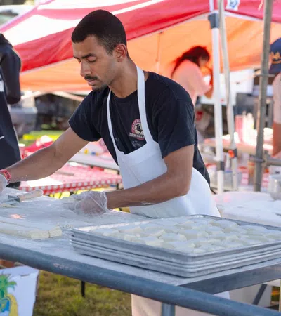Man wearing apron preparing dough at outdoor food stall with trays of dough and tents in background
