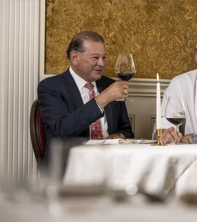 Two men smiling and toasting with red wine in an elegant dining room with gold wallpaper and exposed brick.