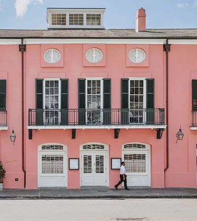 Pink historic building with black shutters, white doors, balconies, and a person walking on the sidewalk.