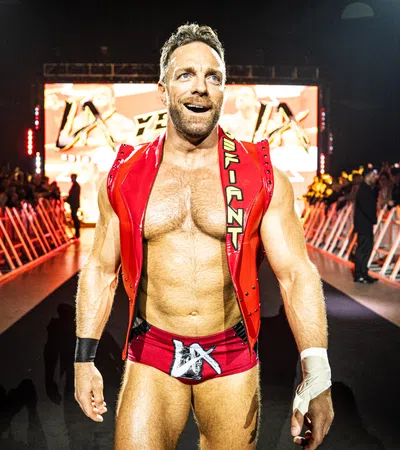 Muscular male wrestler in red trunks and vest walking confidently toward ring with audience and lights behind him
