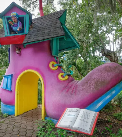 Large whimsical pink boot-shaped playhouse with blue roof and yellow door in a park setting