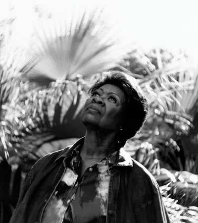 Black and white photo of a woman looking up surrounded by large palm leaves in soft light.