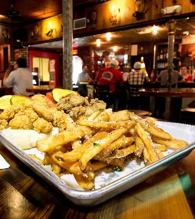 Tray of fried seafood and seasoned fries next to a tall glass of Cooter Brown's beer inside a lively bar.