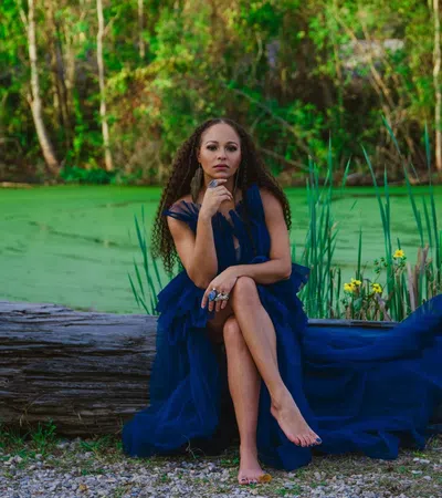 Woman in a flowing navy blue dress sitting barefoot on a log by a green pond surrounded by trees and plants.