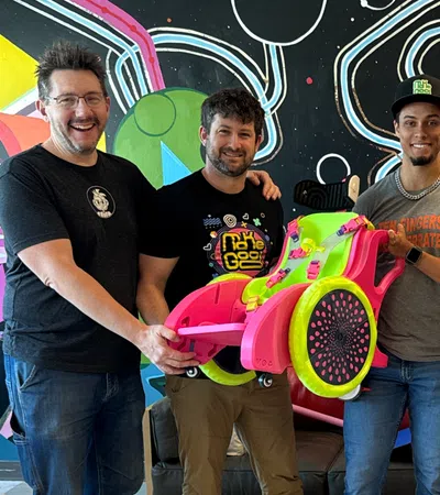 Group of five people smiling indoors with colorful wall art, holding a bright pink and green custom wheelchair.